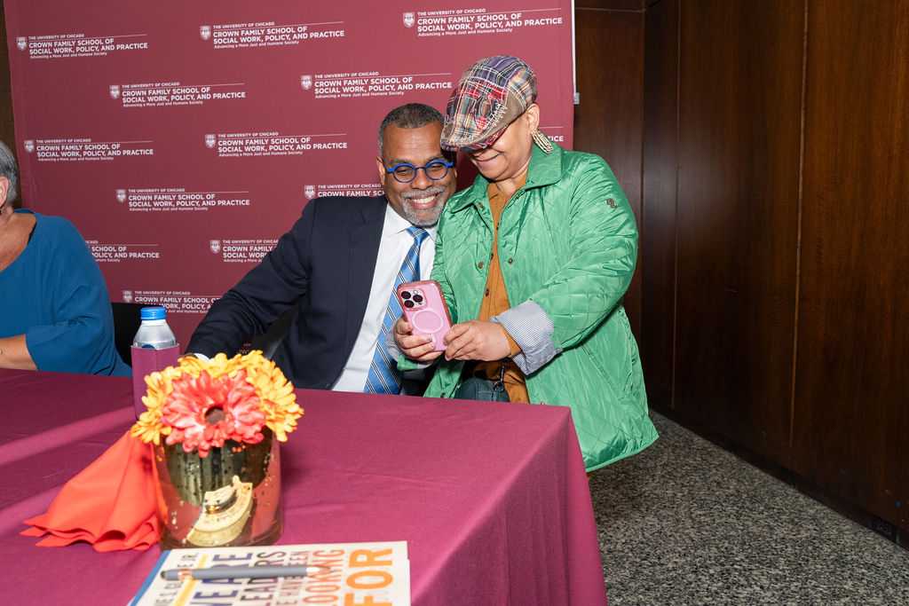 woman in a green coat standing taking a selfie with a man in a suit sitting in front of a step and repeat banner