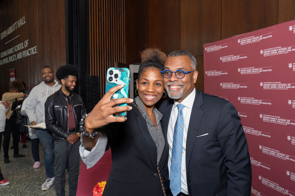 woman in black holding a phone taking a selfie next to a man in a black suit with a tie