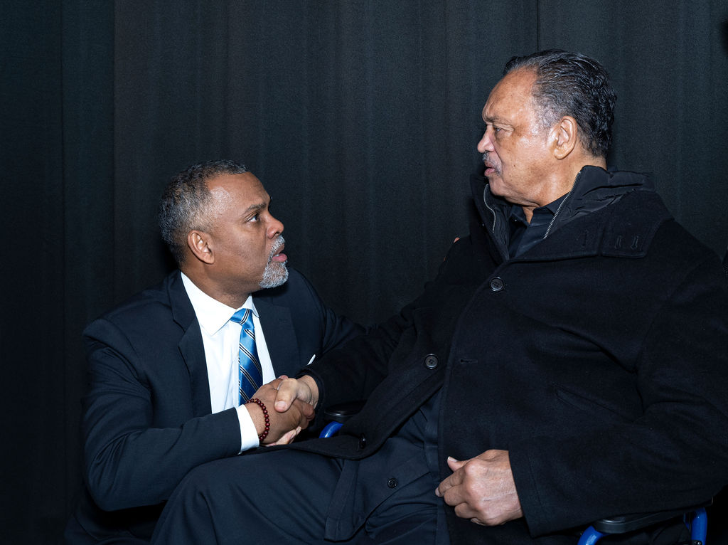 man kneeling next to man in a chair to shake his hand