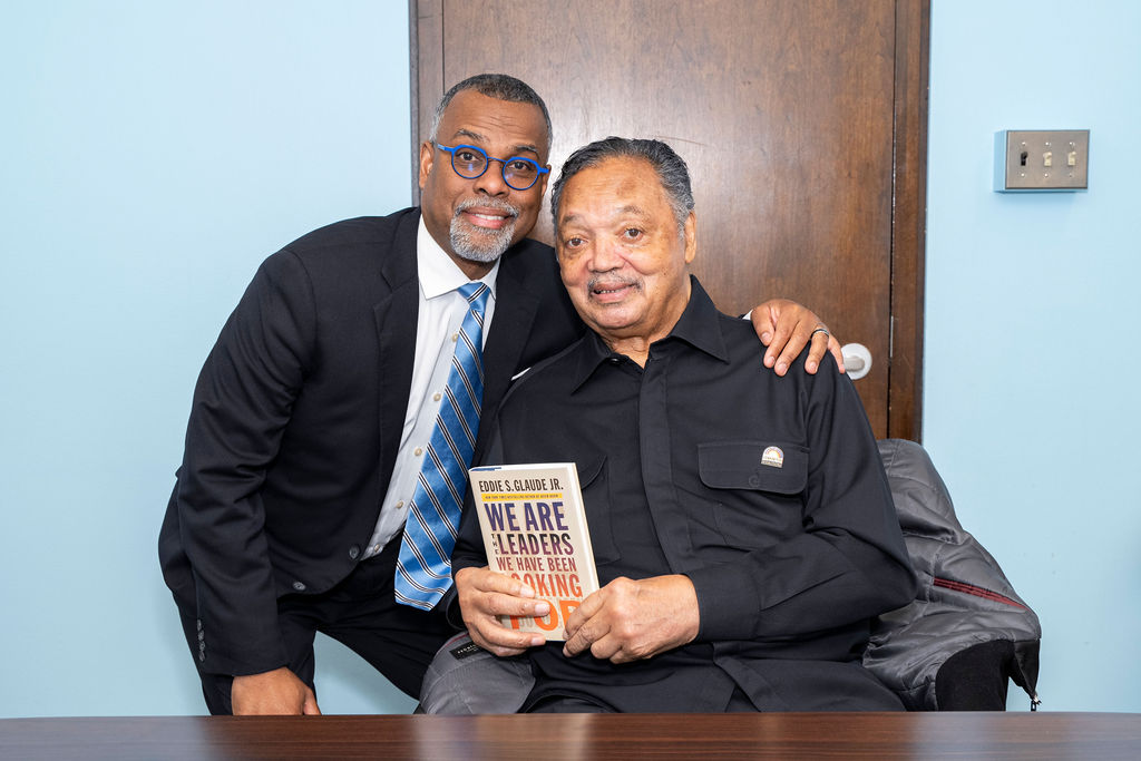 man in suit smiling next to man in black holding a book