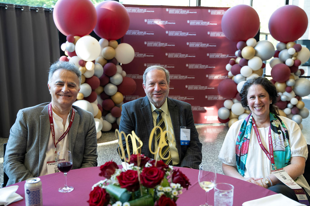 Three people sit at a table facing the camera with balloons in the background