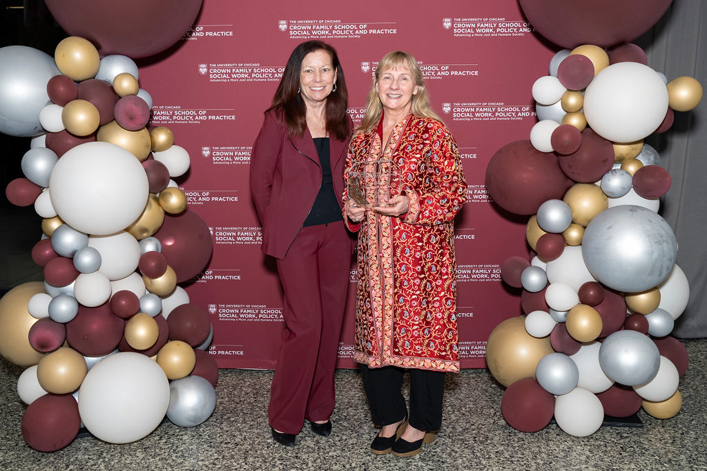 woman in maroon pant suit smiling next to a woman in a colorful blazer holding an award in front of a step and repeat with balloons