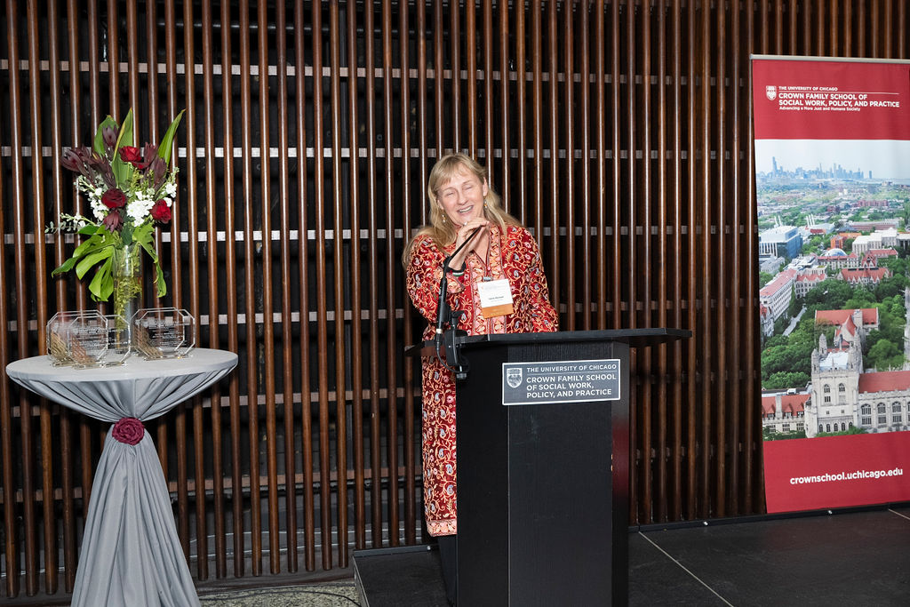 woman in colorful garb stands at podium speaking next to a table with flowers and awards