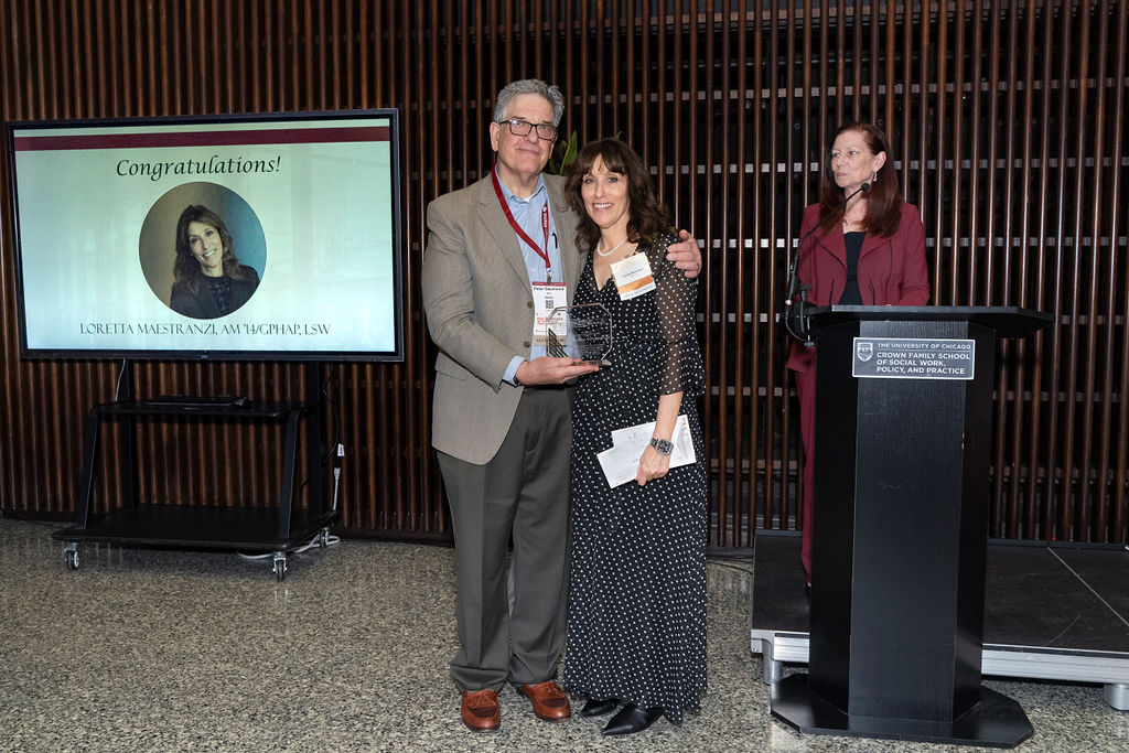 man in grey suit jacket with blue shirt holding an award next to a woman in a black and white polka dot dress smiling while a woman in a maroon suit stands at the podium smiling