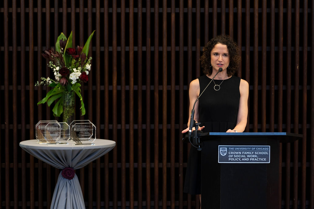 woman in black dress stand at podium speaking next to a table with awards