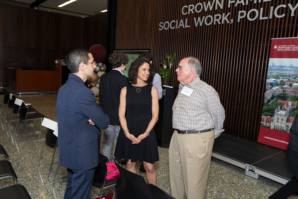 woman in black dress with circle necklace smiling and chatting with two men one in a blue suit and another with a brown checkered shirt with khakis