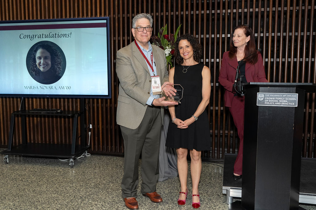 man in grey suit with blue shirt stands holding an award next to a woman in black dress smiling next to a woman at a podium smiling