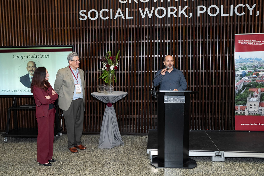 Man in blue shirt standing at podium speaking while woman in maroon and man in grey suit stands near smiling