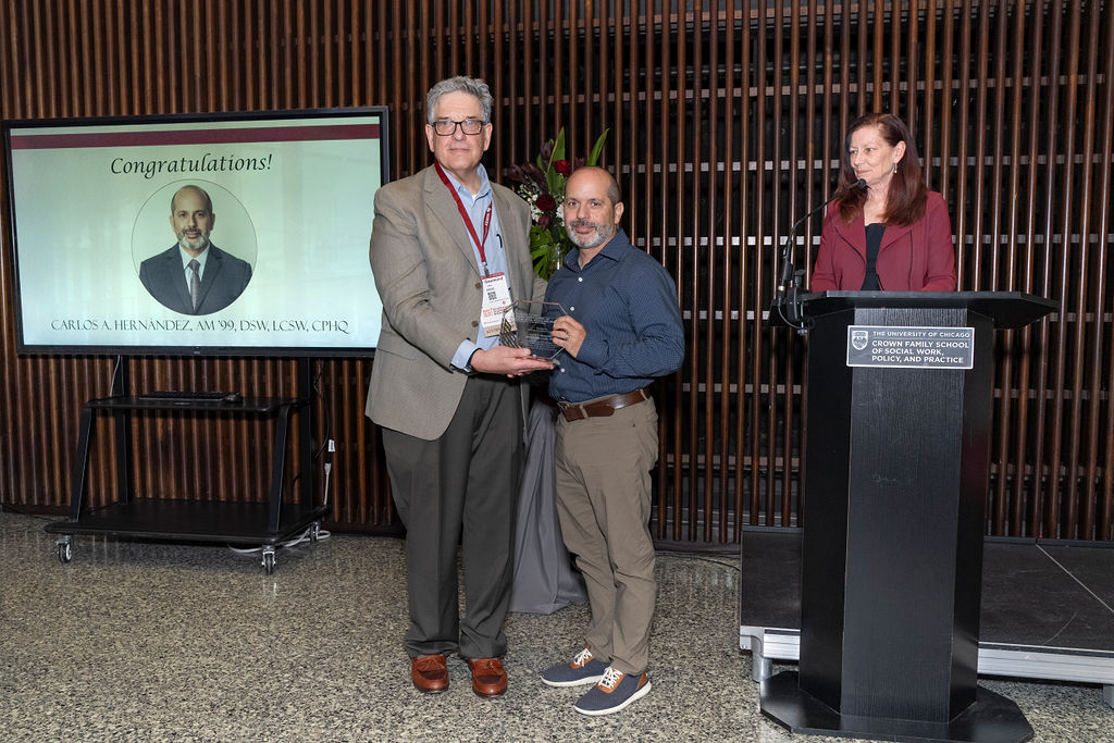 man in grey suit jacket holding an award next to a man in a blue shirt and khakis as a woman in maroon stands at a podium smiling