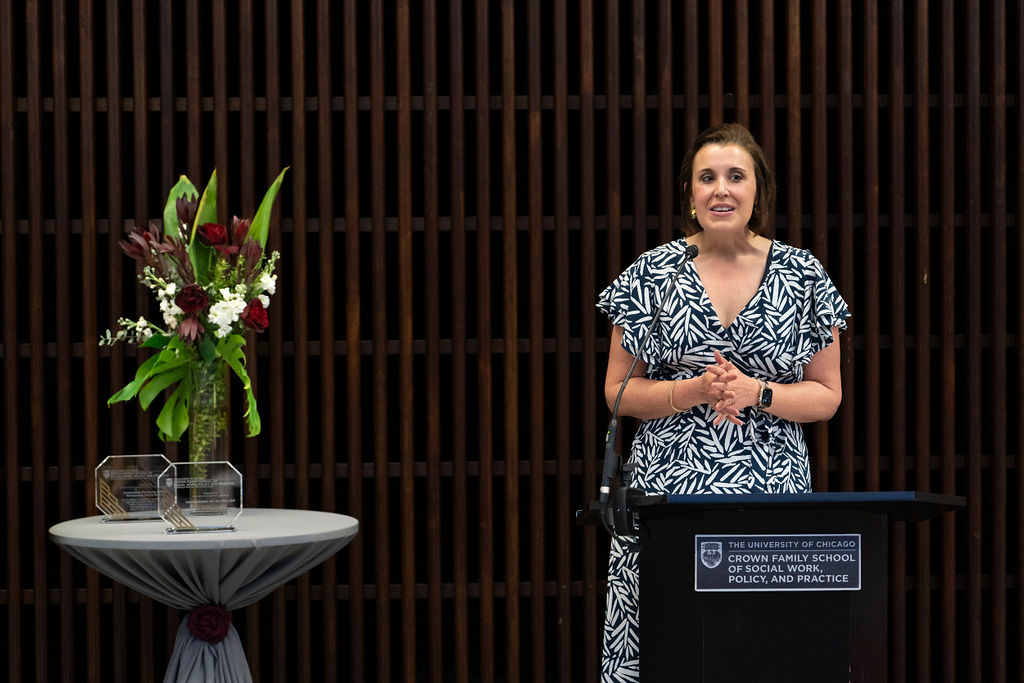 woman in blue and white dress stand at podium speaking next to a table with flower an awards