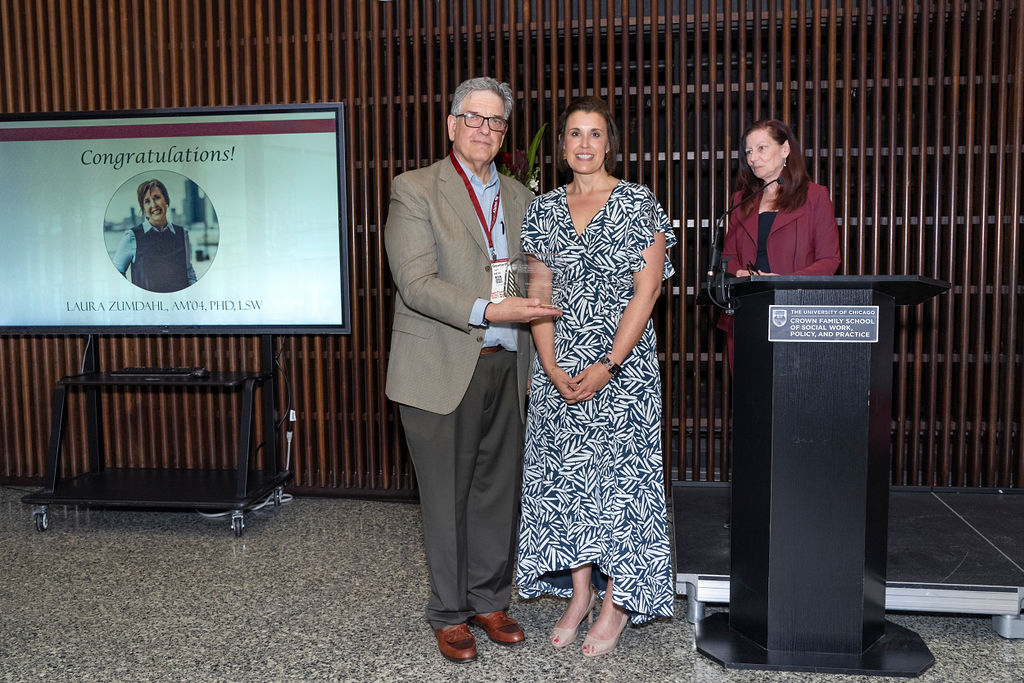 man in grey suit holding an award next to a woman in a blue and white dress while a woman in maroon stands at the podium smiling