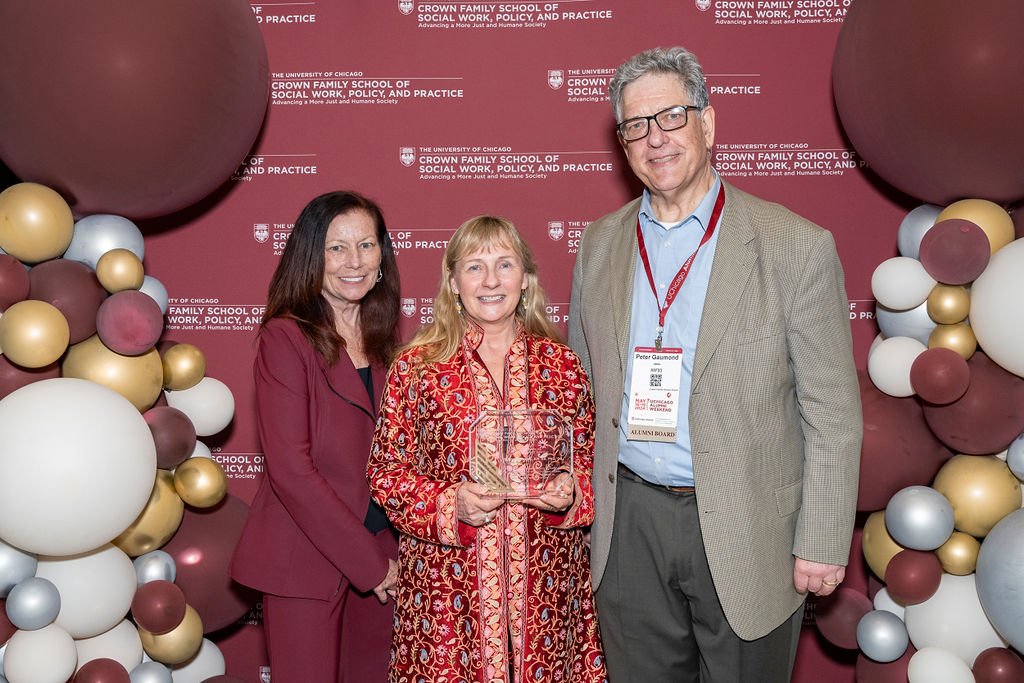 woman in maroon standing next to a woman in colorful garb holding an award next to a man in a grey suit jacket in front of a step and repeat with balloons smiling