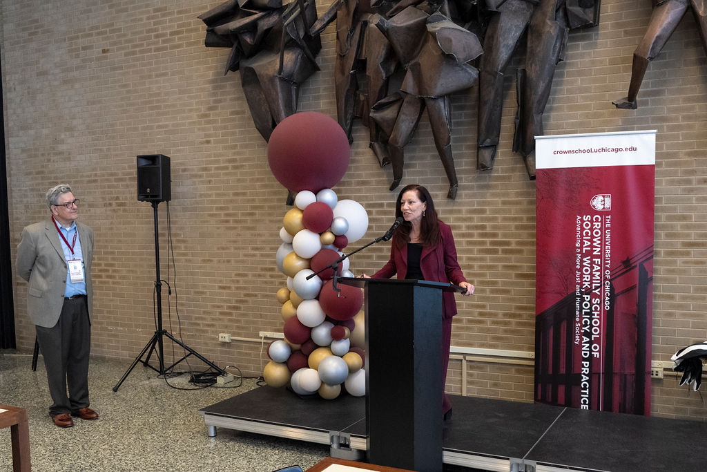 Woman in maroon pants suit with black shirt speaking at a podium with balloons behind her.