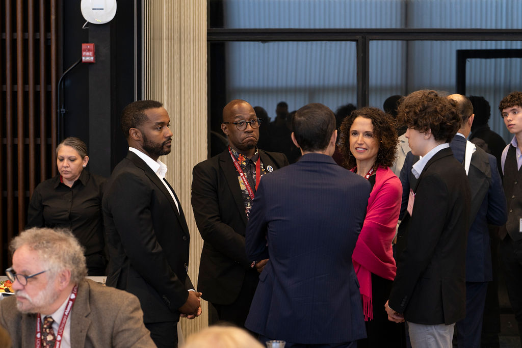 man in black suit and white shirt chatting with man in black suit multi colored shirt standing next to a woman in pink shawl with a young man in black suit jacket and male in a blue suit 