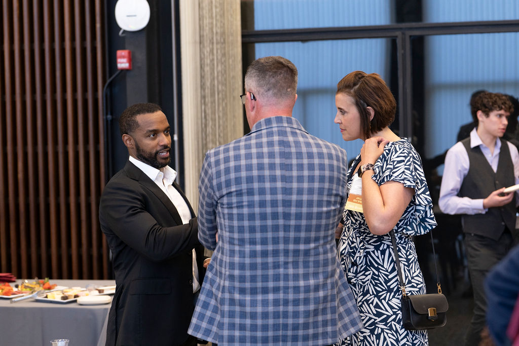 man in black suit with white shirt chatting with male in blue checked suit and woman in a blue and white dress