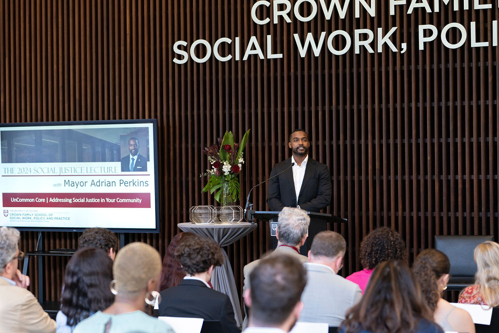 Male in a black shirt with white shirt standing at a podium speaking to a an audience next to a monitor with his name and picture