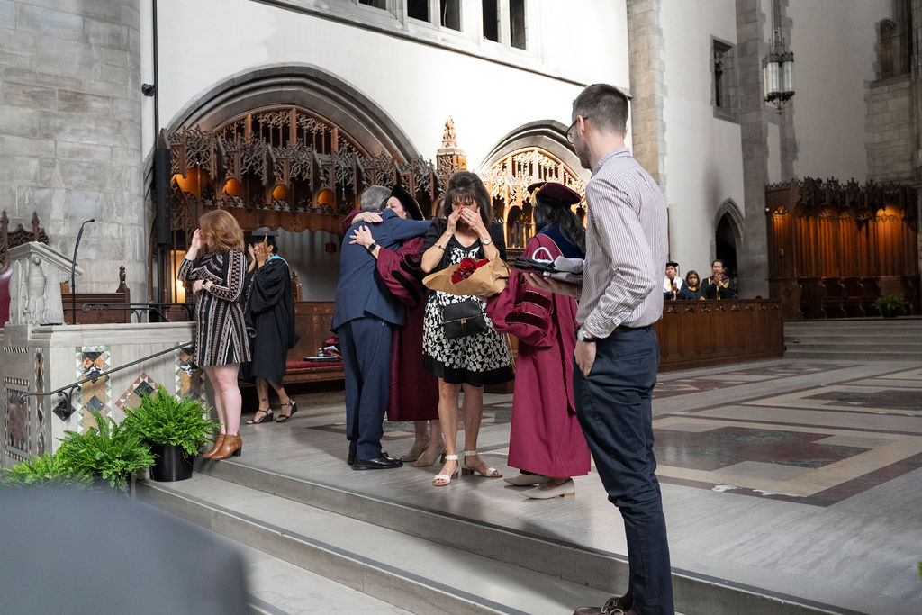 woman in a maroon robe hug a man in a suit as a woman in black and white wipe here tearful eyes