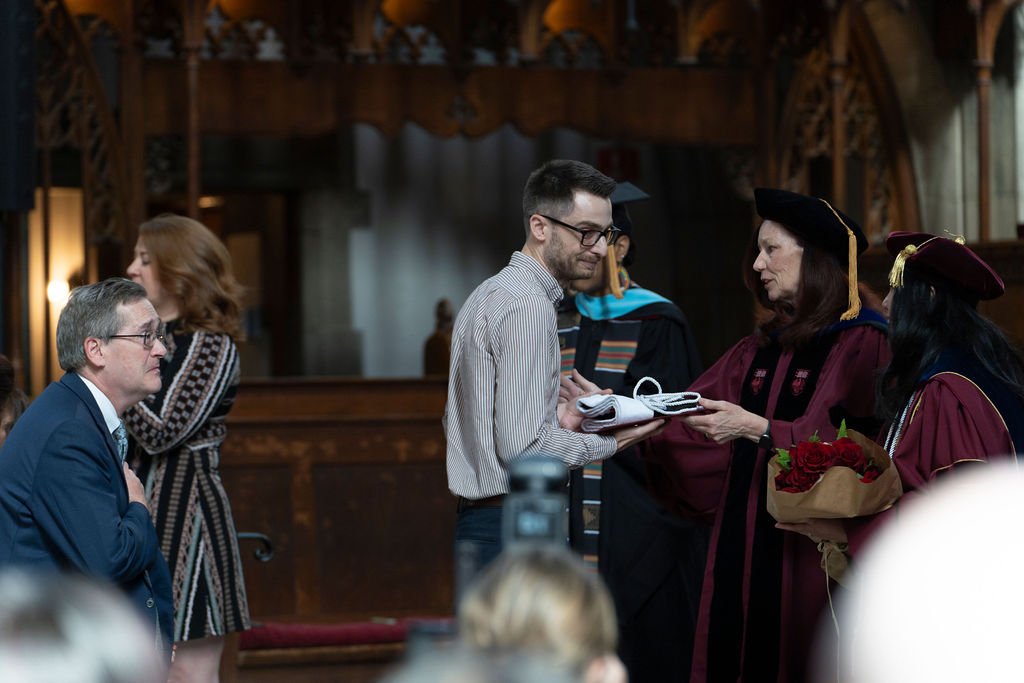 a man in striped shirt receive a folded hood from a woman in a maroon robe