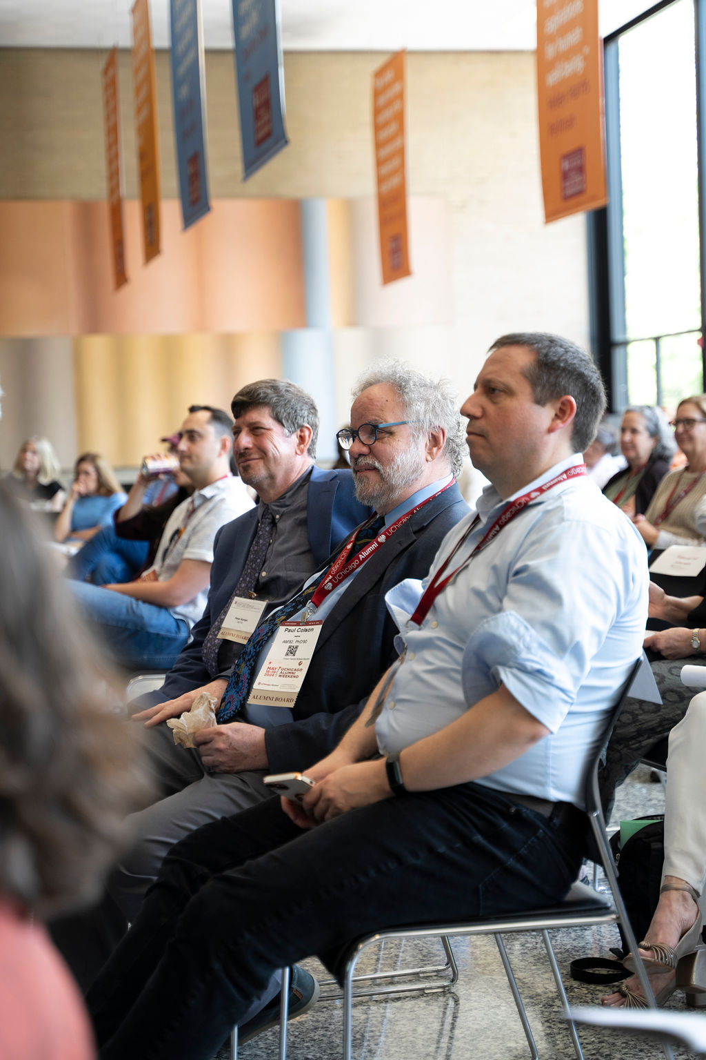 Three male presenting persons sit in an audience listening to a speaker