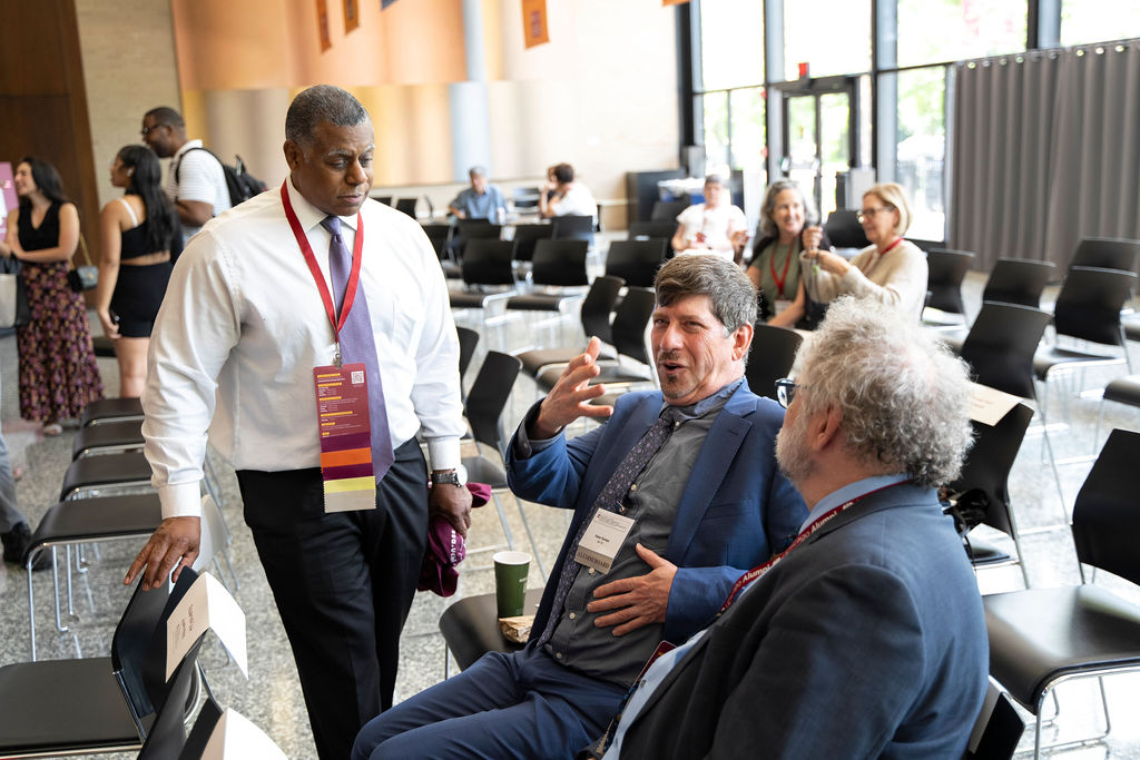 One male presenting person stands near chairs speaking to two seated male presenting persons