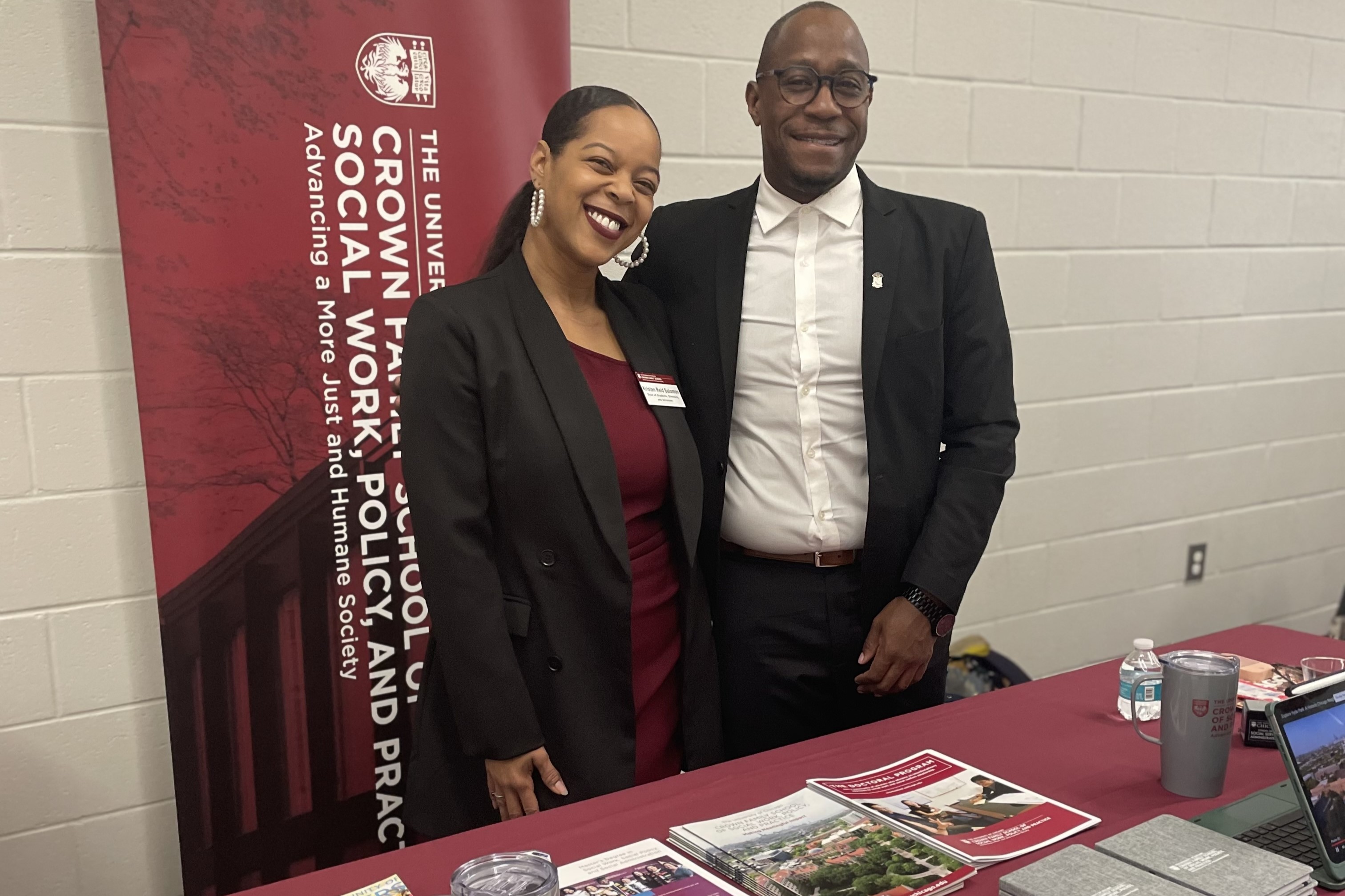 woman in black jacket standing next to man in black jacket with with shirt smiling while at a table for recruitment