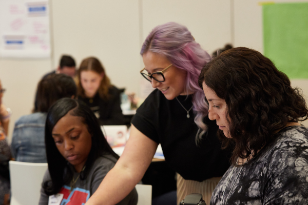 women with pink hair leaning over guiding two other women at a table