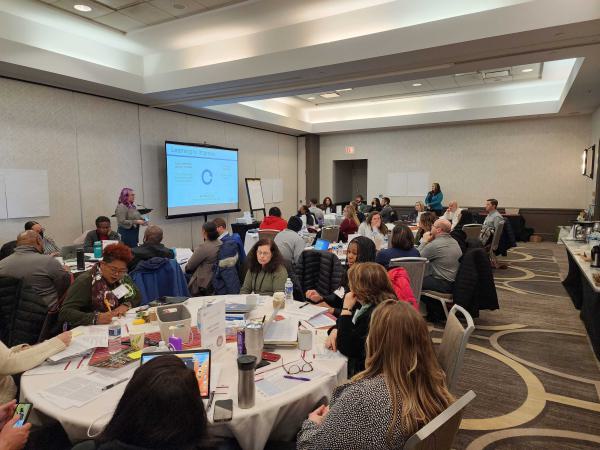 A room full of people listening at round tables to a speaker