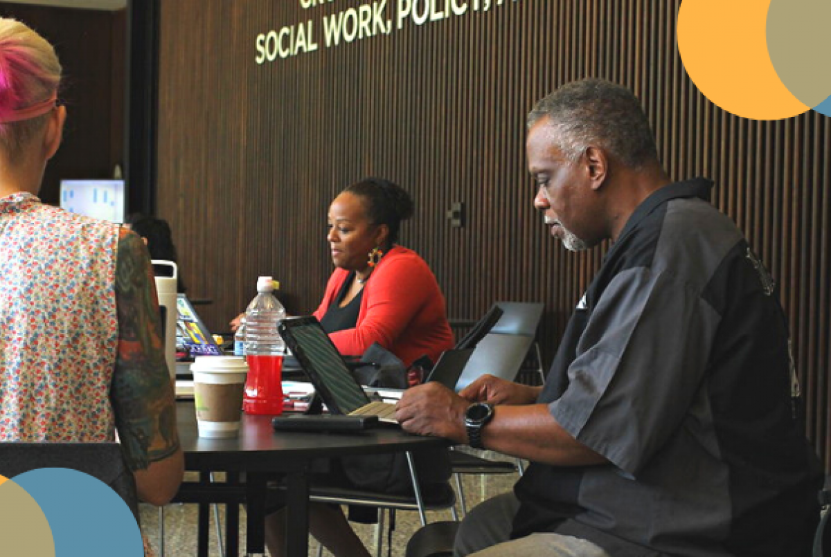 image of a man in a black shirt sitting at a table on his laptop with two other people. 