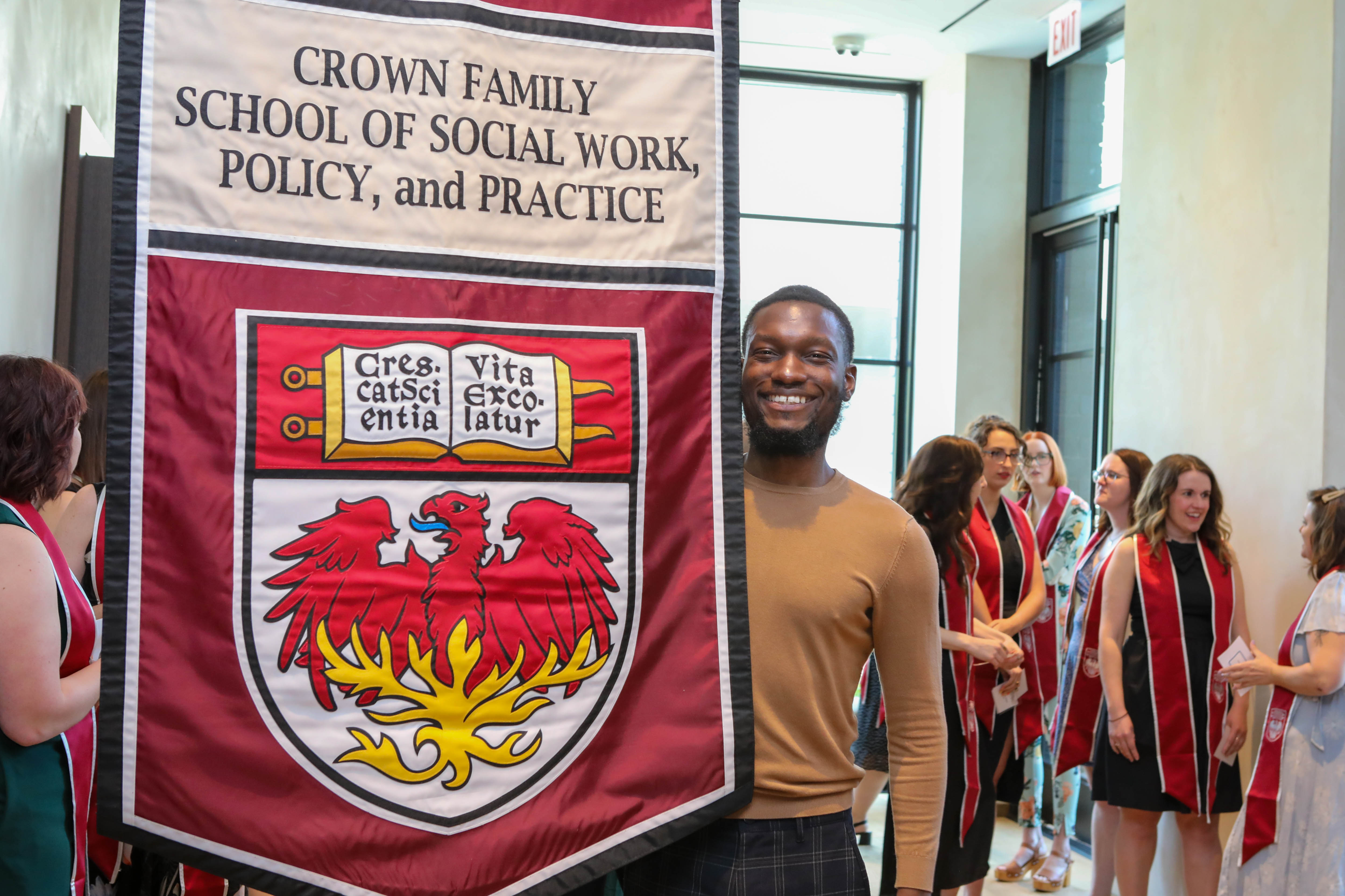 male standing in a gold sweater holding a school flag smiling