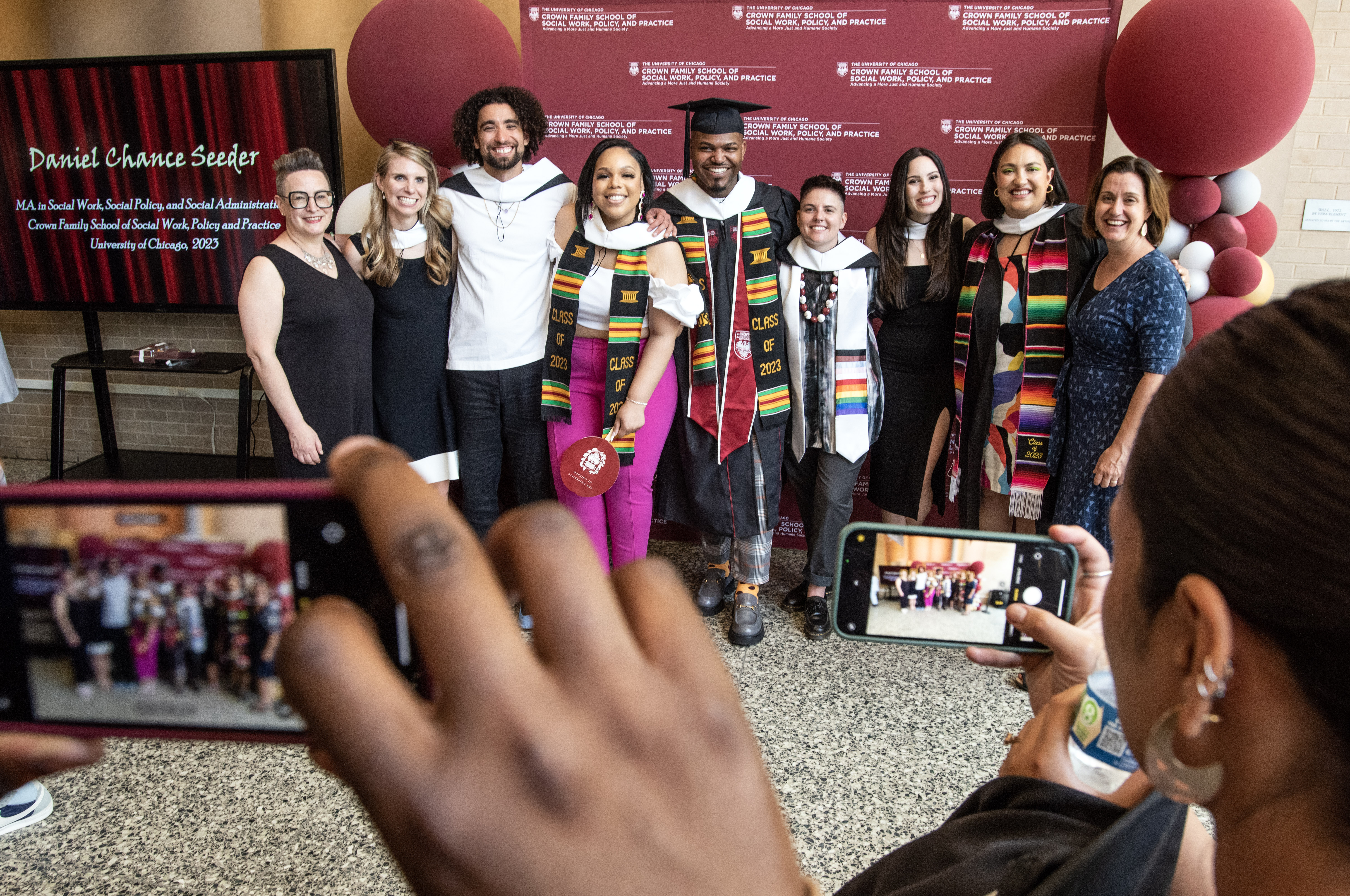 a group of students in graduation robes smiling in front of a maroon step and repeat with balloons