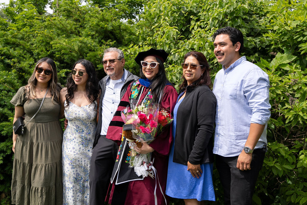 A PhD graduate stands holding flowers with family members on either side of her all wearing sunglasses on the lawn