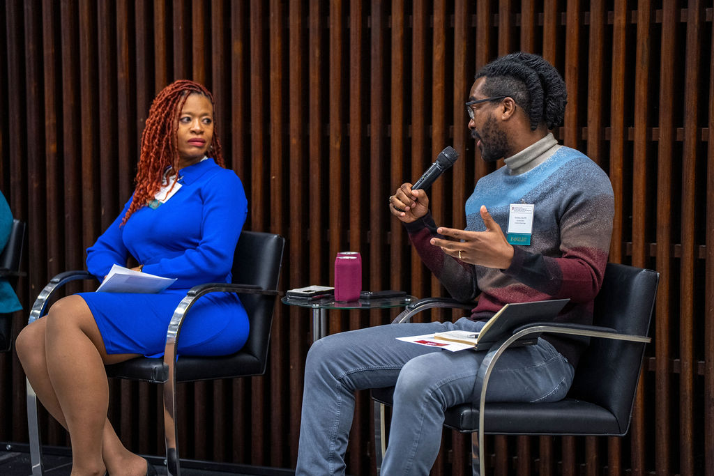 Black woman with red hair in blue dress looking at black man in sweater and jeans speaking with microphone.