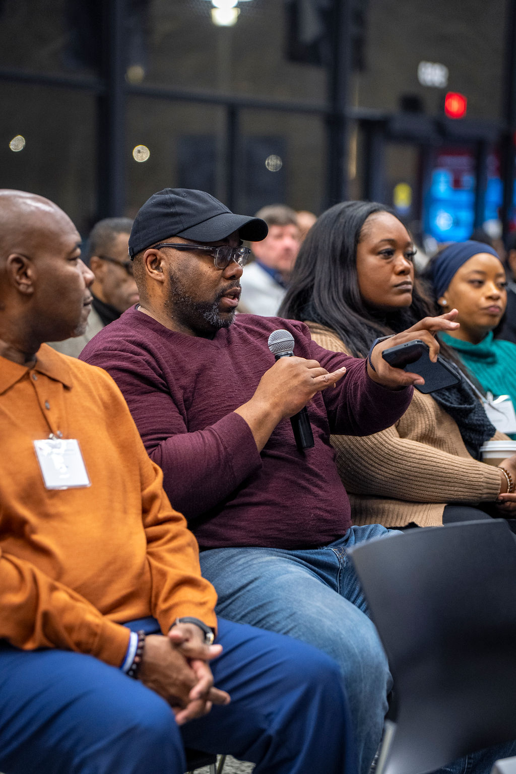 Man in maroon sweater is holding a microphone. Man next to him in orange sweater looking closely at speaker.