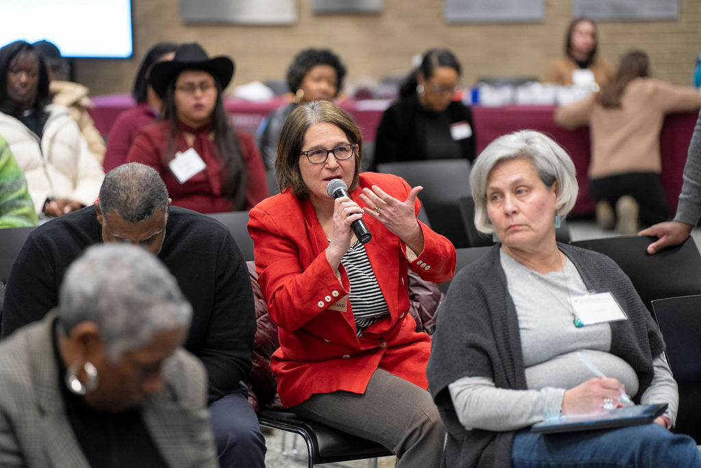 Groiup pof seated people, woman in the middle in red blazer is holding a microphone making a motion with her arm. Woman Infront is in a cardigan and is looking to the side.