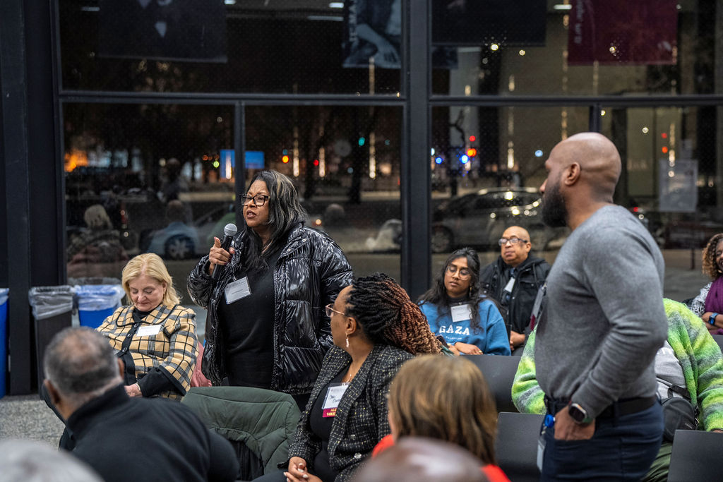 A woman is standing amongst a group is seated. The woman is holding a microphone and is looking attentively.