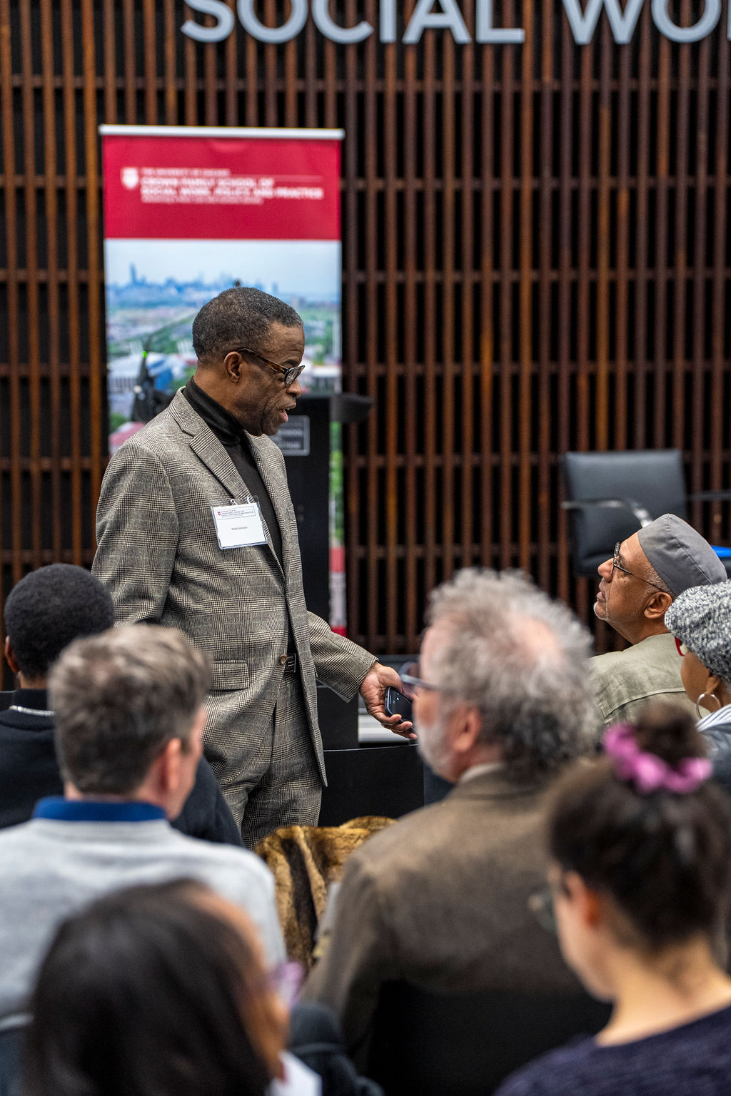 People sitting down, a man in a grey blazer is standing and talking to an indivdual