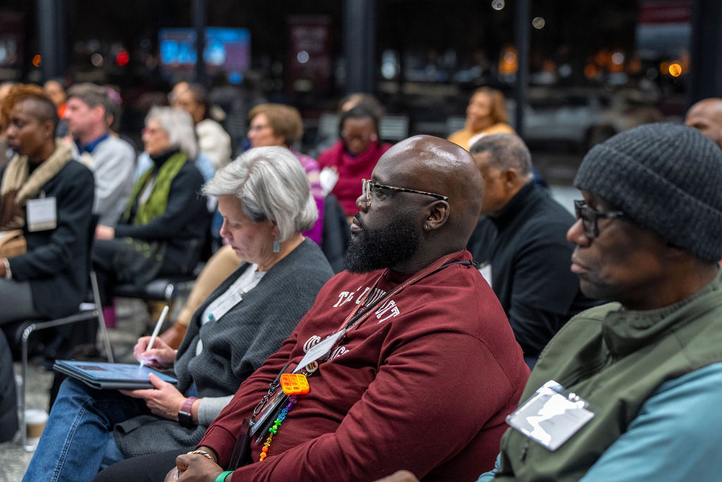 Black man with a beard and glasses wearing a red shirt sitting in a chair in the crowd.