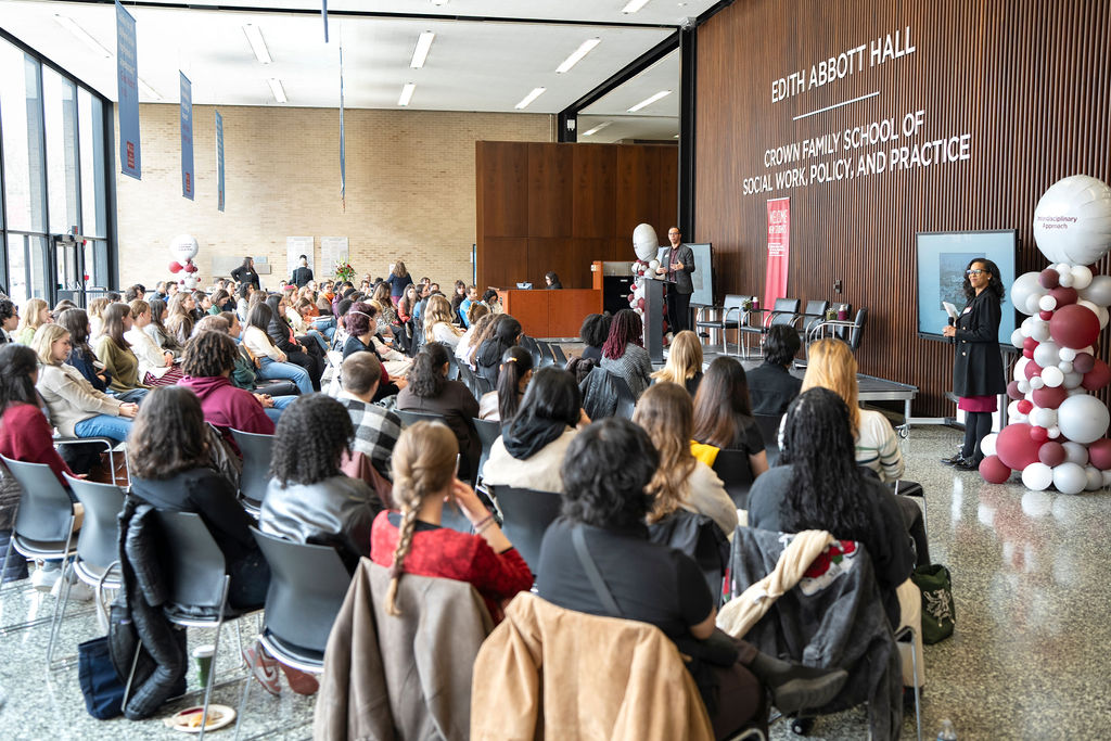 Audience facing the speaker, a man in a dark grey suit jacket, maroon and grey balloons to each side of the stage.