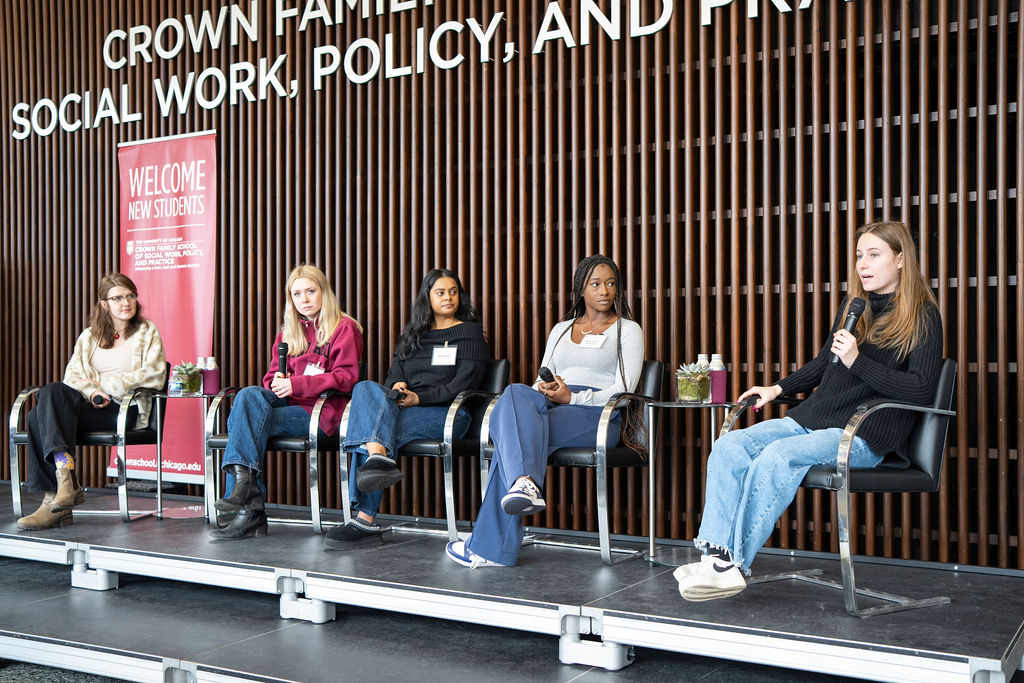 Five woman seated in a line on a stage, focused on woman with black sweater as they hold a microphone.
