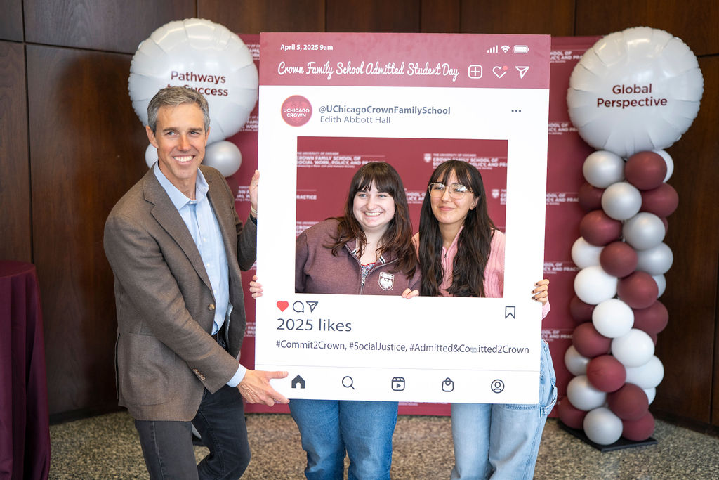 Man in brown suit jacket smiling as he holds up the social media photo prop for a woman in crew neck and a woman in a pink sweater