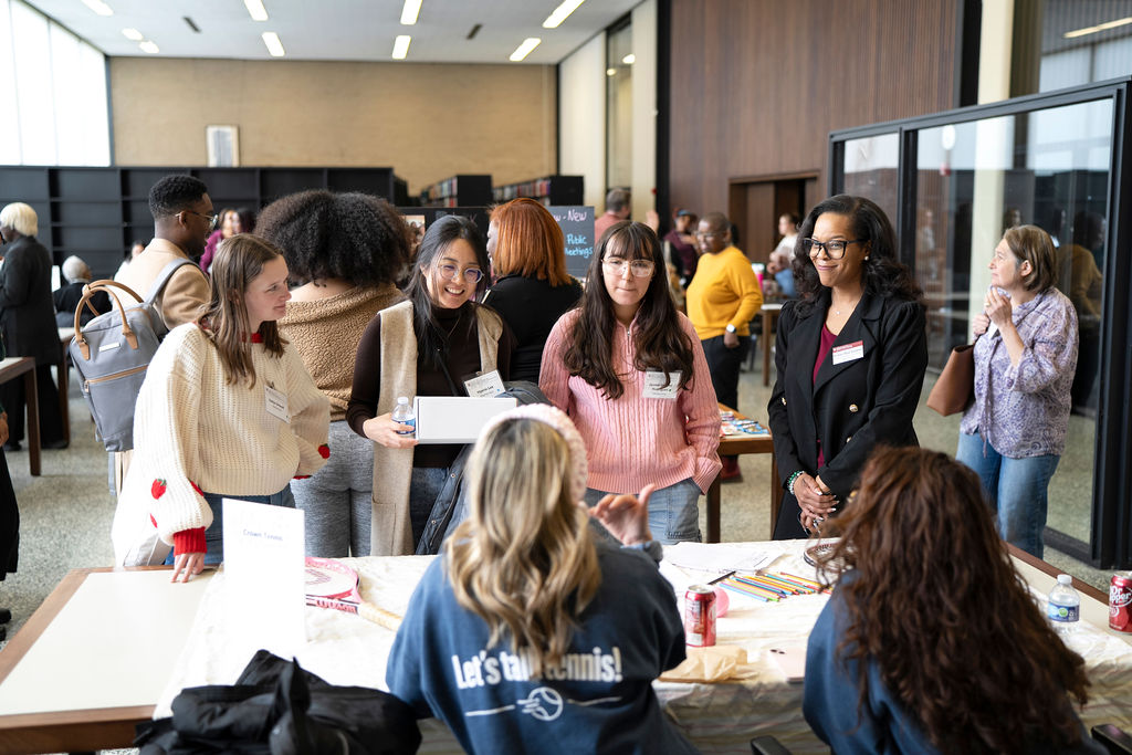 Three women standing in front of table with two women in blue shirts talking