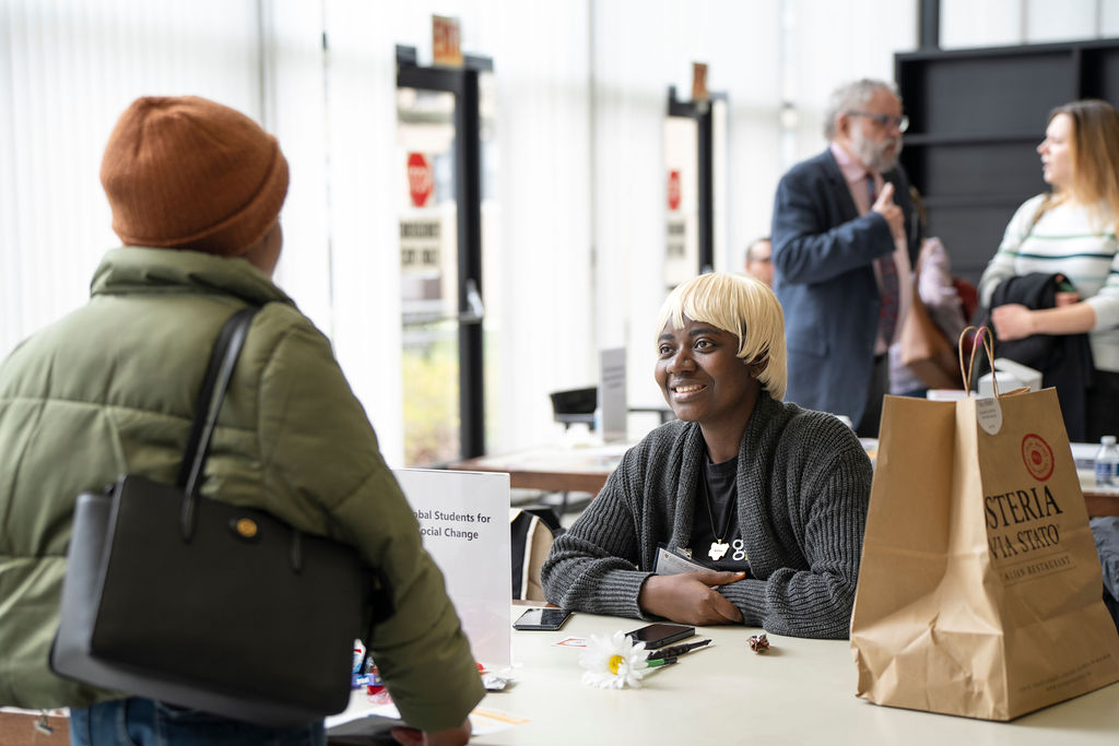 Black woman with blonde hair sitting behind table talking to black woman in green jacket and beanie