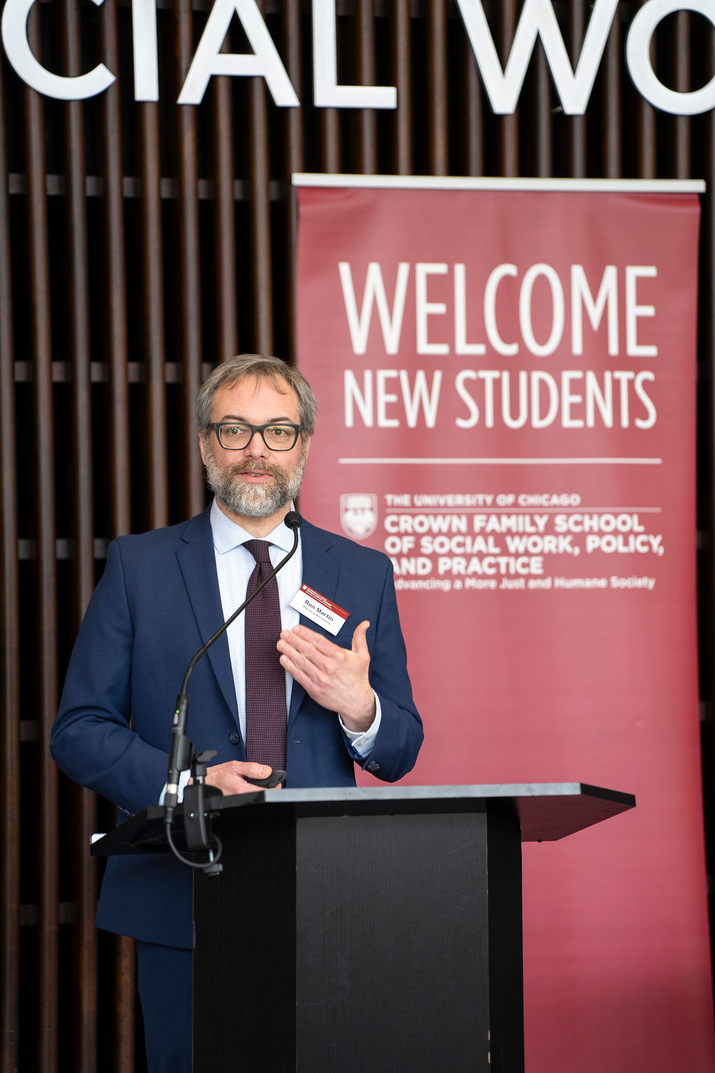 Man in blue suite wearing glasses standing behind podium, the banner behind him reads, "Welcome New Students".