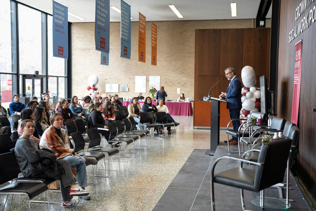 Suited man is standing behind a podium onstage as audience is seated right in front of the stage.