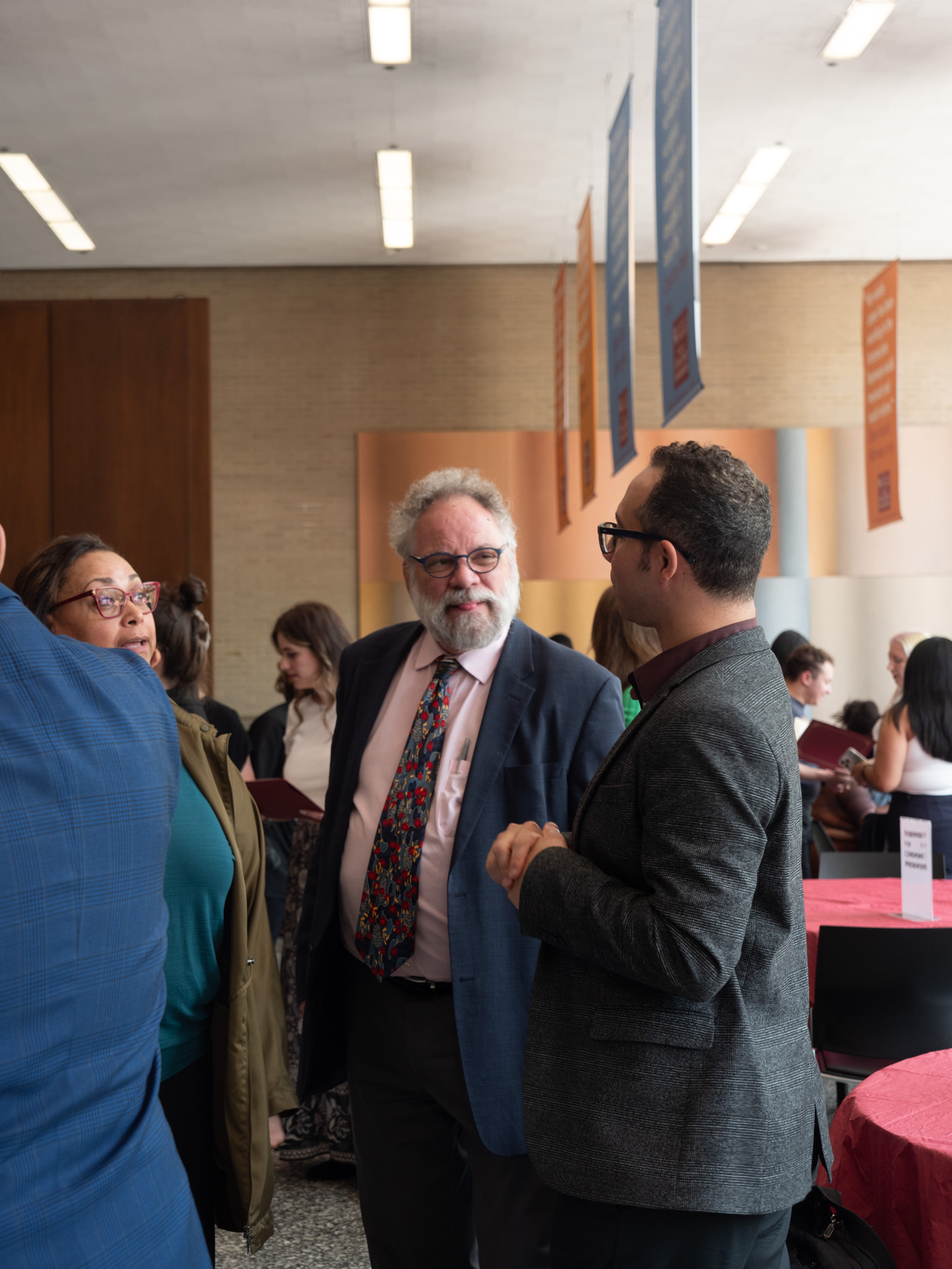 Man in grey suit facing man in pink shirt and blue suit jacket talking to woman in glasses