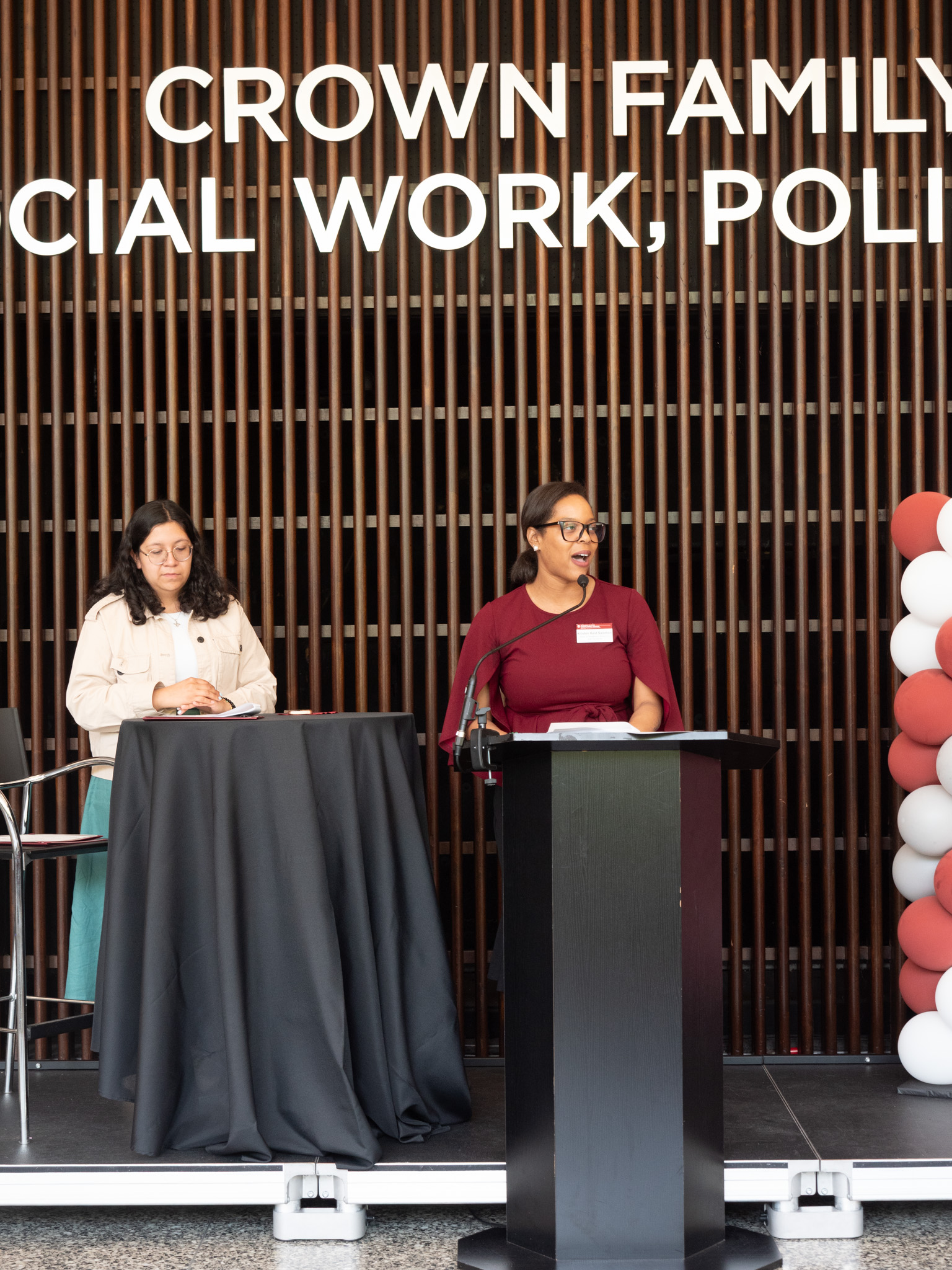 Woman in red looks of the ide from behind a podium a second woman with cream colored cardigan stands a few feet behind woman in red.