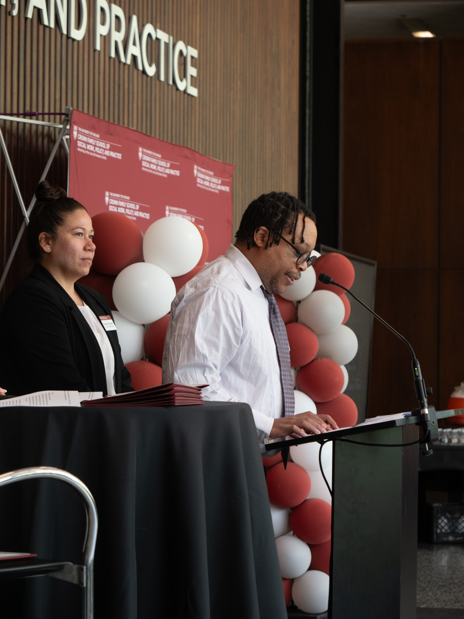 Man in white formal shirt and tie looks down at podium as woman in black cardigan stands a few feet from him.