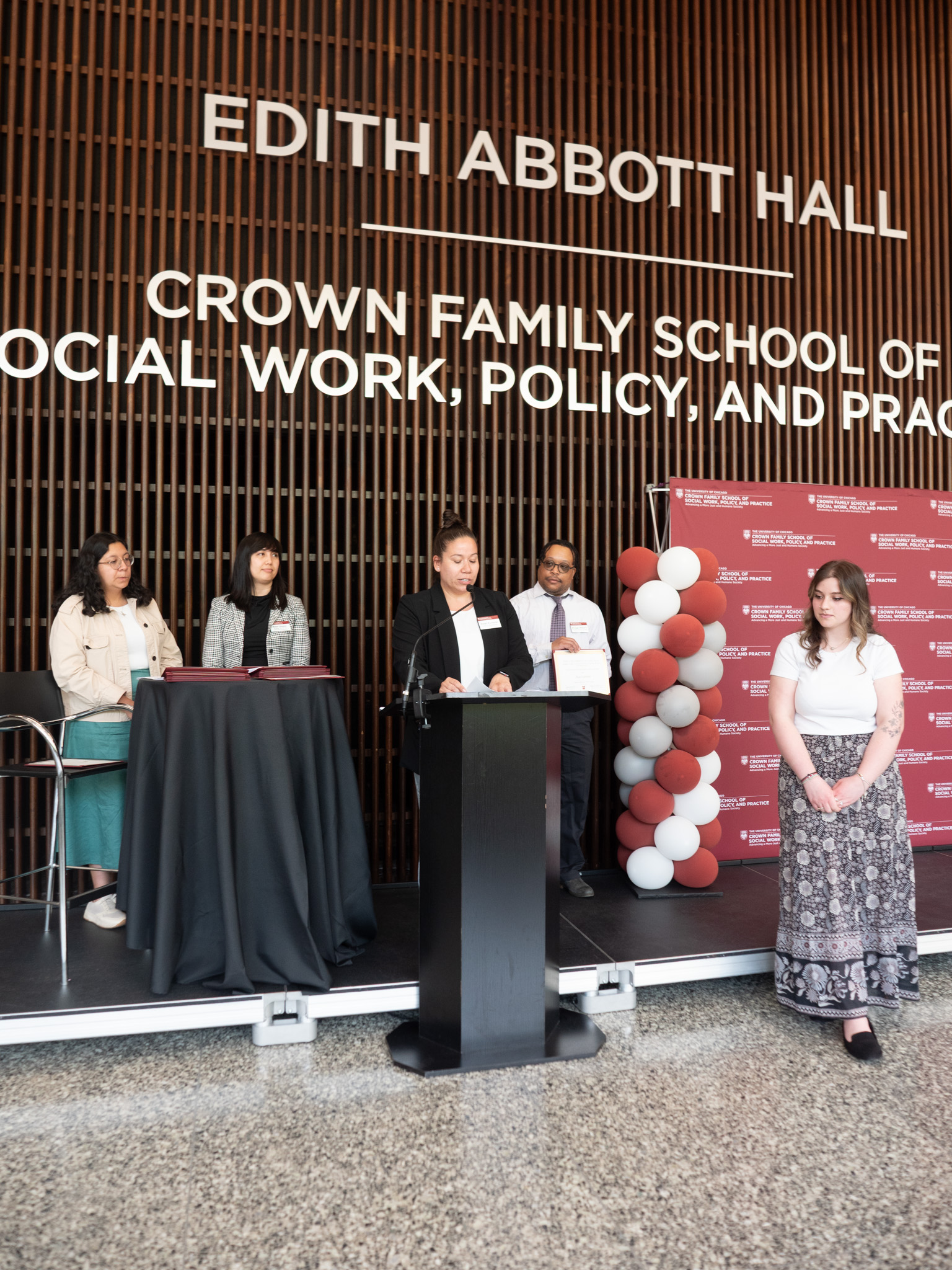 Student awardee wearing white shirt and grey floral skirt standing next to podium in front of stage