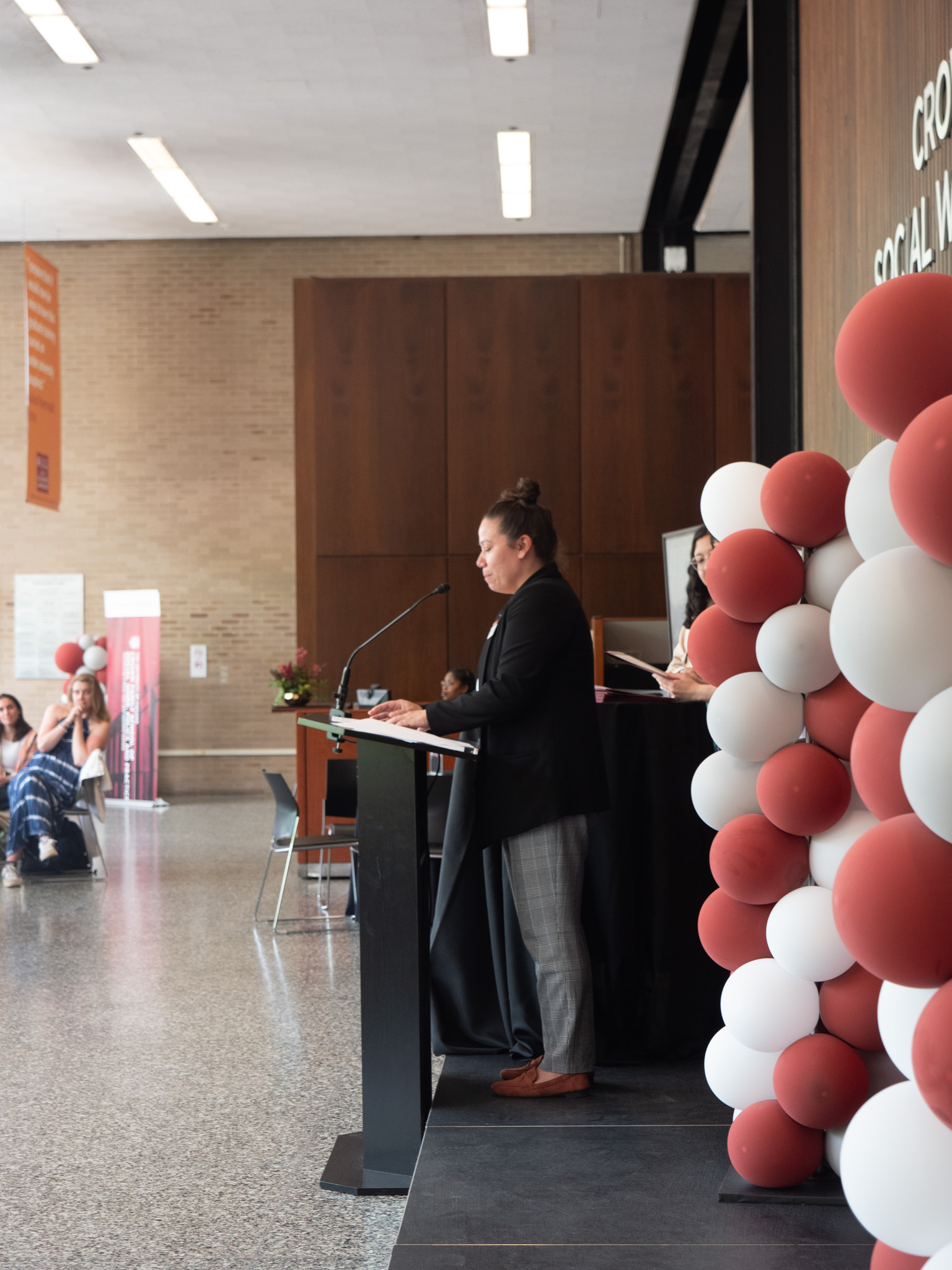 Woman with hair in high bum and black cardigan looking down at podium as they stand in front of a few balloon decorated pillars