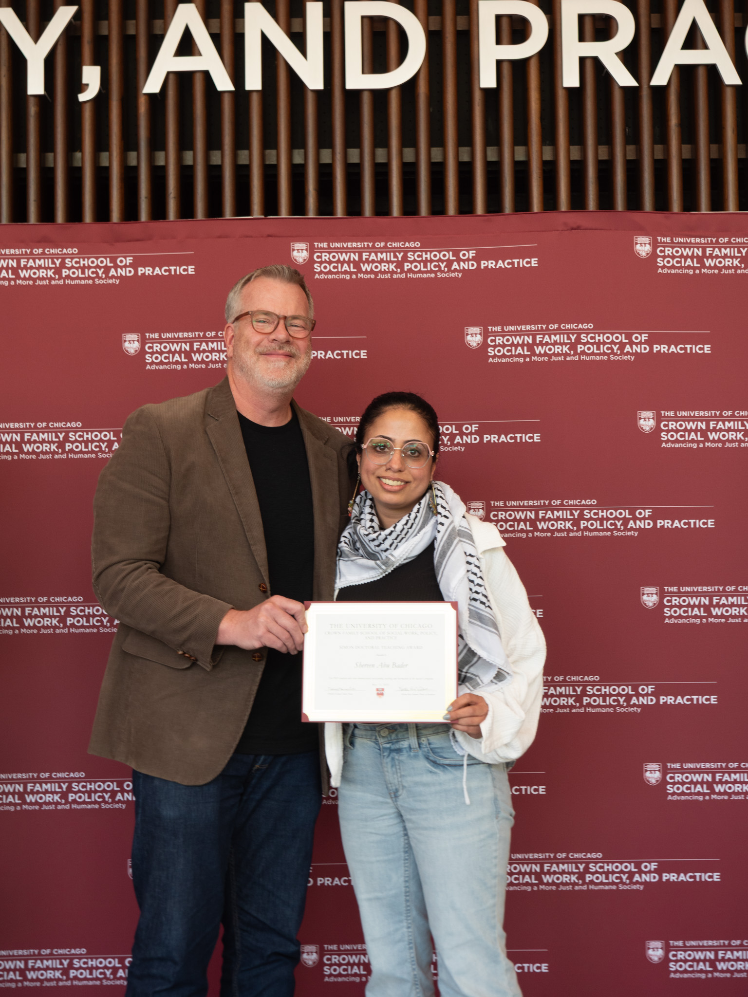 Student awardee wearing white and black scarf and blue jeans taking picture with man in brown suit jacket in front of backdrop